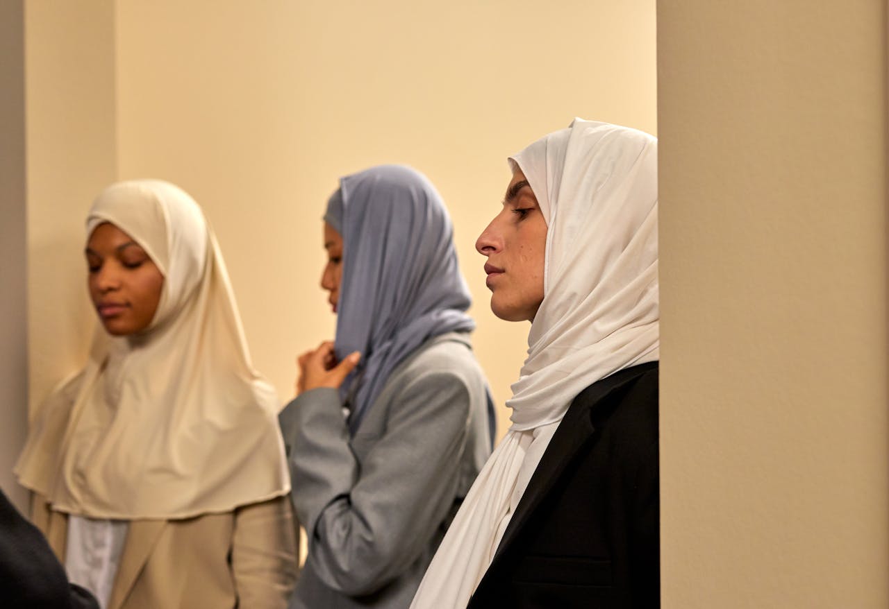 Three women in hijabs standing indoors with eyes closed, conveying diversity and contemplation.
