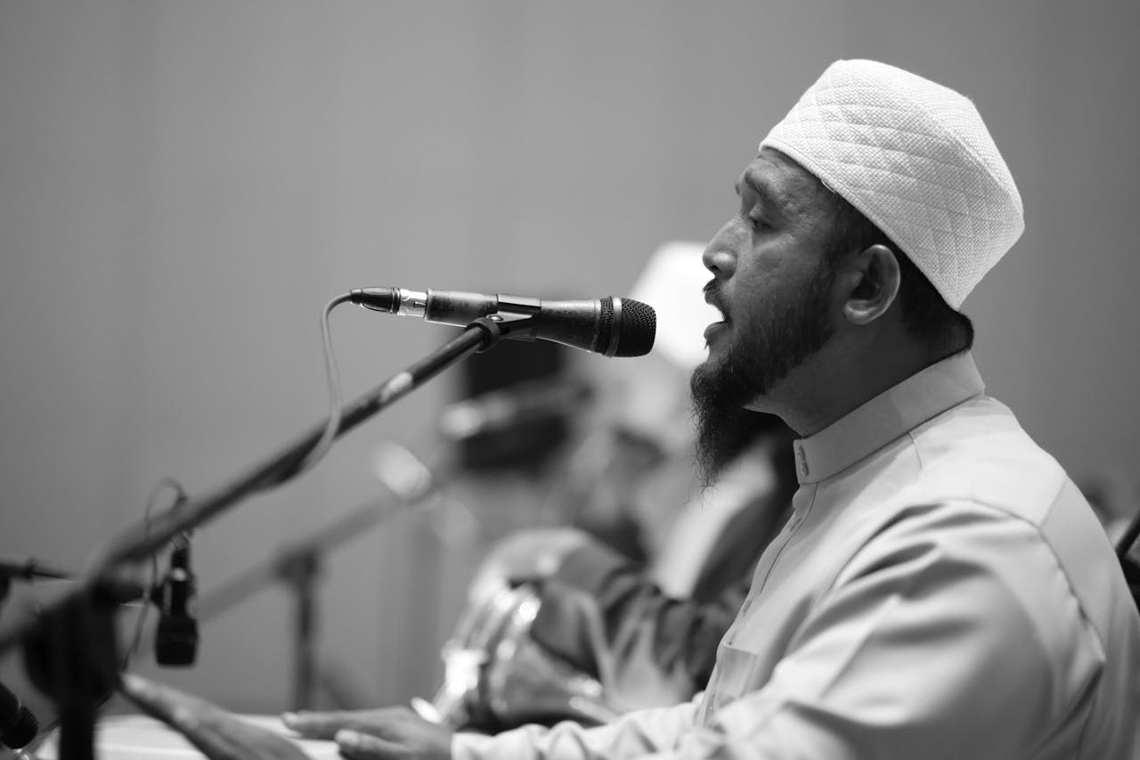 Side view of a man in traditional attire speaking into a microphone in a monochrome setting.